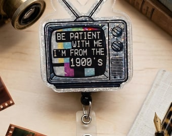 May include: A glittery acrylic badge reel shaped like a vintage television set with two antennas. The screen displays the text BE PATIENT WITH ME I'M FROM THE 1900'S over a colourful test pattern. The device includes a retractable cord and a metal clip.