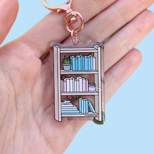 Könnte beinhalten: Ein durchsichtiger Acryl-Schlüsselanhänger mit einem Miniatur-Bücherregal mit einer Pflanze, Büchern und einem Hai. Das Bücherregal ist braun mit weißen Regalbrettern und die Bücher sind in Blautönen, Rosa und Weiß. Der Schlüsselanhänger hat einen roségoldenen herzförmigen Verschluss.