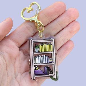 May include: A clear acrylic keychain featuring a bookshelf with three shelves. The top shelf has a small plant and yellow books. The middle shelf has white books and a small jar. The bottom shelf has purple and black books with a bee on top. The keychain has a gold metal clasp.