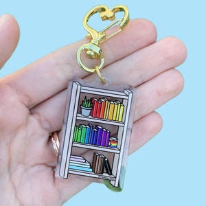 以下が含まれることがあります： カラフルな本、鉢植えの植物、虹色のカップが置かれた本棚のデザインのアクリルキーホルダー。ゴールドカラーのハート型クラスプと小さなリングが付いています。「Scrambled Pegs」の文字が下部にあります。