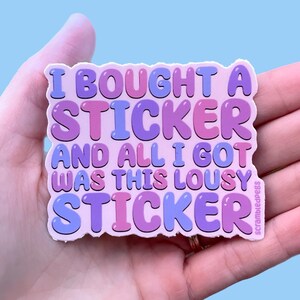 Op de afbeelding: Een sticker met de tekst "I BOUGHT A STICKER AND ALL I GOT WAS THIS LOUSY STICKER" in bubbelletters. De sticker heeft een lichtroze achtergrond met paarse en blauwe tekst. De sticker wordt in een hand gehouden.