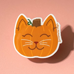May include: A die-cut sticker featuring a smiling cat face shaped like a pumpkin. The orange pumpkin cat has white whiskers, a brown stem, and the text "Scrambled Pegs" on the side. The sticker is on a pink background.