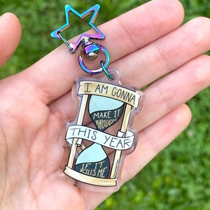 May include: A clear acrylic keychain with a gold hourglass design. The hourglass is filled with blue and black sand and has the text "I am gonna make it through this year if it kills me". The keychain is attached to a rainbow star-shaped metal ring.