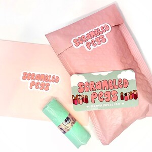 May include: An overhead shot of a package with the text "Scrambled Pegs" on a pink envelope, a pink bubble mailer, and a business card. A small, wrapped item is also visible.