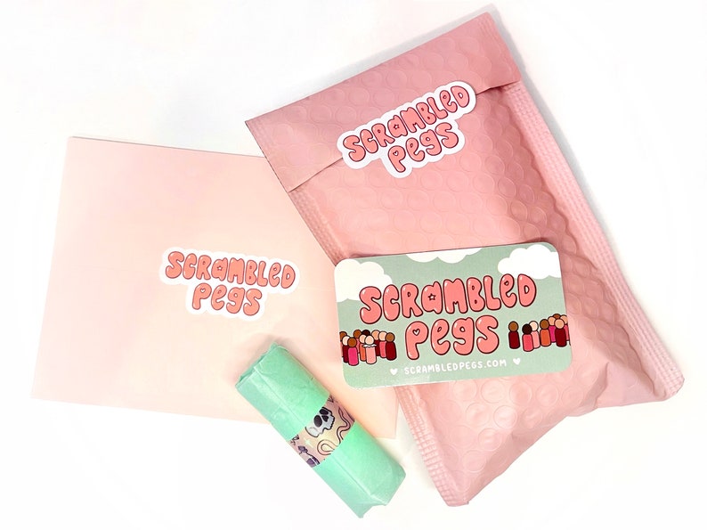 May include: An overhead shot of a package with the text "Scrambled Pegs" on a pink envelope, a pink bubble mailer, and a business card. A small, wrapped item is also visible.