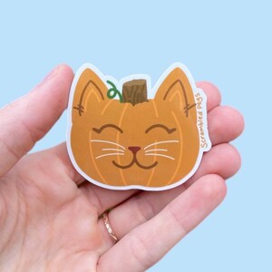 May include: A pumpkin-shaped sticker with a cat face design. The sticker is orange with a brown stem and green vine. The cat face has white whiskers and a smiling mouth. The text "Scrambled Pegs" is on the right side.