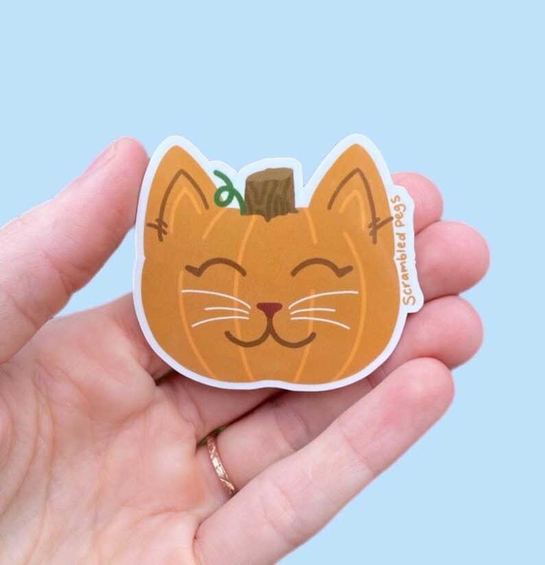 May include: A pumpkin-shaped sticker with a cat face design. The sticker is orange with a brown stem and green vine. The cat face has white whiskers and a smiling mouth. The text "Scrambled Pegs" is on the right side.