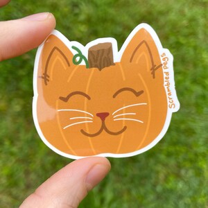 May include: A die-cut sticker featuring a smiling cat face designed as a pumpkin. The orange pumpkin cat has a brown stem, green vine, and white whiskers. The sticker has the text "Scrambled Pegs" on the right side.