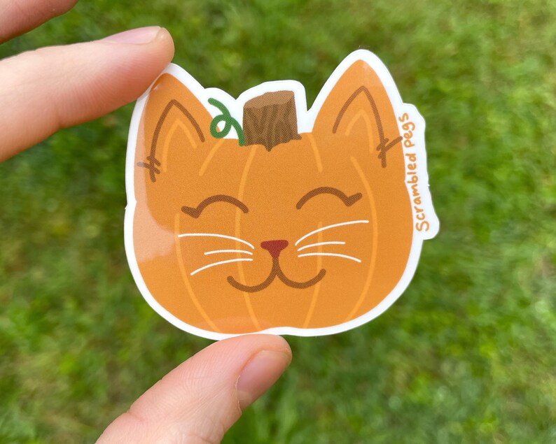 May include: A die-cut sticker featuring a smiling cat face designed as a pumpkin. The orange pumpkin cat has a brown stem, green vine, and white whiskers. The sticker has the text "Scrambled Pegs" on the right side.