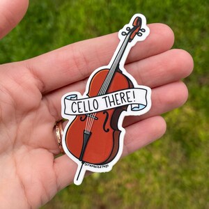 K&ouml;nnte beinhalten: Ein wei&szlig;er Aufkleber mit einer Cello-Illustration und dem Text "Cello There!" in Schwarz. Das Cello ist braun mit einem hellbraunen Hintergrund. Der Aufkleber befindet sich auf einer Hand.