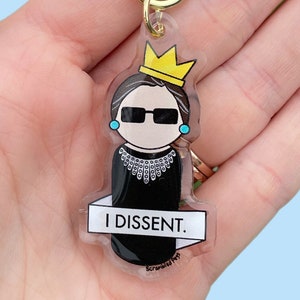 May include: A clear acrylic keychain featuring a cartoon illustration of Ruth Bader Ginsburg wearing a black robe, a yellow crown, and sunglasses. The keychain has the text "I DISSENT." printed on a white banner.