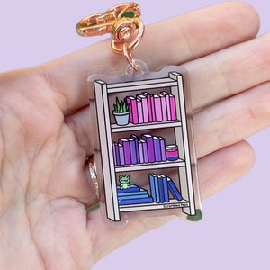 Op de afbeelding: Een transparante acryl sleutelhanger in de vorm van een boekenkast met een klein kikkerfiguurtje, een plant in een pot en kleurrijke boeken. De sleutelhanger heeft een roségouden metalen sluiting en de tekst "Scrambled Eggs" onderaan.