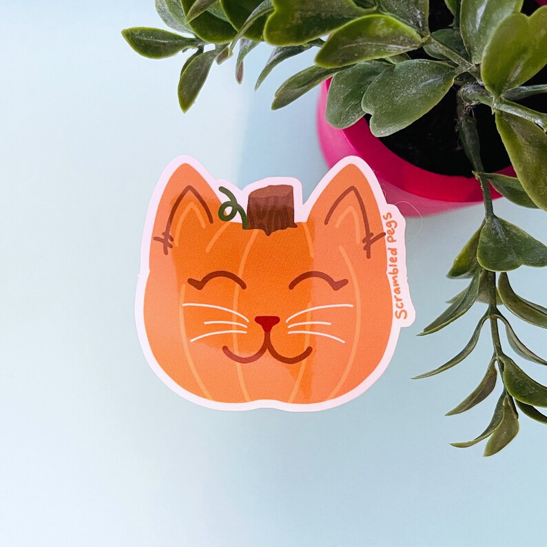 May include: A sticker featuring a smiling cat face designed as a pumpkin. The orange pumpkin cat has a brown stem, stitched ears, and white whiskers. The sticker has the text "Scrambled Pegs" on the right side.