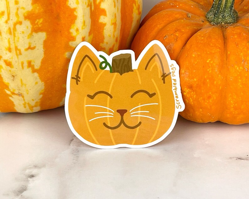 May include: A die-cut sticker featuring a smiling cat face designed as a pumpkin. The sticker is orange with a brown stem and green vine. The text "Scrambled Pegs" is printed on the sticker.