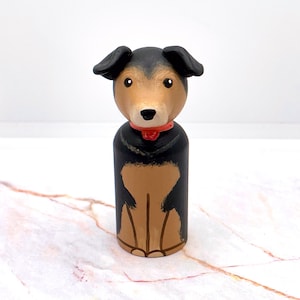 Puede incluir: Figura de perro de madera con pelaje negro y canela, collar rojo y orejas caídas. El perro está pintado con un acabado liso y está sentado erguido. El objeto es decorativo.