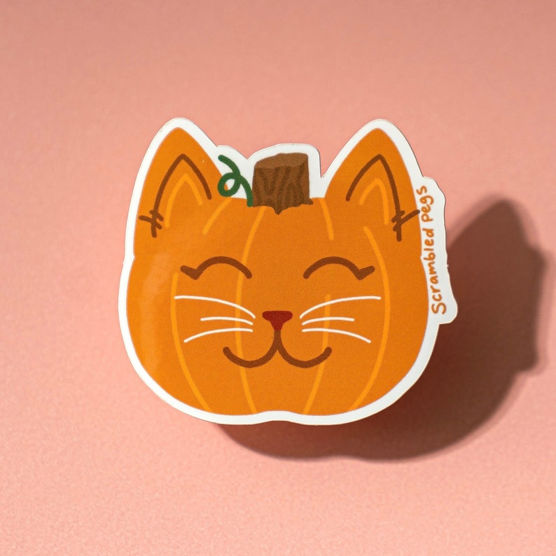 May include: A die-cut sticker featuring a smiling cat face shaped like a pumpkin. The orange pumpkin cat has white whiskers, a brown stem, and the text "Scrambled Pegs" on the side. The sticker is on a pink background.