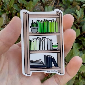 Puede incluir: Una pegatina que representa una pequeña estantería con tres estantes. Los estantes están llenos de libros en tonos verdes, una pequeña planta en maceta y una vela. La pegatina tiene el texto "Scrambled Pegs" en la parte inferior.