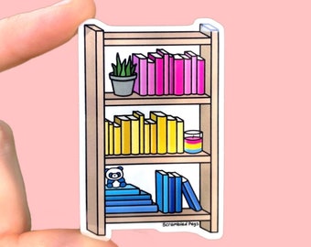 May include: A sticker of a wooden bookshelf with three shelves. The top shelf has a potted plant and a row of pink and purple books. The middle shelf has a row of yellow books and a small candle with a pansexual pride flag design. The bottom shelf has a blue panda plushie and a stack of blue books. The sticker has the text "Scrambled Pegs" at the bottom.
