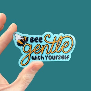 Könnte beinhalten: Ein rechteckiger Aufkleber mit türkisfarbenem Hintergrund und den Worten "bee gentle with yourself" in einer verspielten Schriftart. Eine Cartoon-Biene befindet sich links. Der Aufkleber wird von einer Hand gehalten.