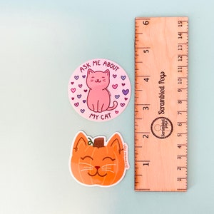 May include: Two stickers on a light blue surface. One is a pink cat with the text "ASK ME ABOUT MY CAT" surrounded by hearts. The other is an orange pumpkin-shaped cat. A wooden ruler is next to the stickers.