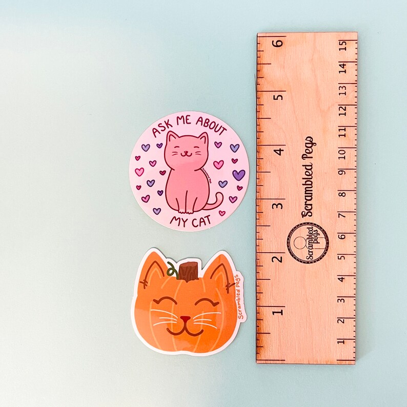 May include: Two stickers on a light blue surface. One is a pink cat with the text "ASK ME ABOUT MY CAT" surrounded by hearts. The other is an orange pumpkin-shaped cat. A wooden ruler is next to the stickers.