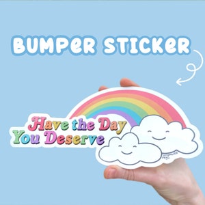 May include: A rainbow sticker with two clouds and the text "Have the Day You Deserve". The sticker is held in a hand against a light blue background.