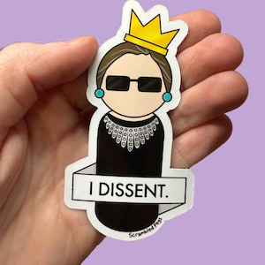 Könnte beinhalten: Ein schwarz-weißer Aufkleber mit einer Karikatur von Ruth Bader Ginsburg, die eine Krone, eine Sonnenbrille und eine schwarze Robe mit weißem Kragen trägt. Der Aufkleber trägt den Text "I DISSENT." und den Text "Scrambled Pegs" unten.