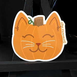 May include: A die-cut sticker featuring a smiling cat face shaped like a pumpkin. The orange pumpkin cat has white whiskers, brown eyes and nose, and a green vine stem. The text "Scrambled Pegs" is on the right side.