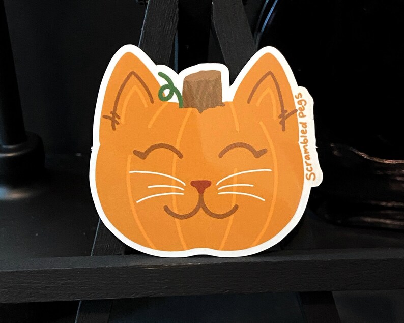 May include: A die-cut sticker featuring a smiling cat face shaped like a pumpkin. The orange pumpkin cat has white whiskers, brown eyes and nose, and a green vine stem. The text "Scrambled Pegs" is on the right side.