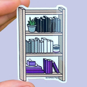 Puede incluir: Una pegatina de una estantería con tres estantes. El estante superior tiene una planta en maceta y una fila de libros negros. El estante del medio tiene una fila de libros grises y una vela pequeña. El estante inferior tiene un pastel morado y una pila de libros morados. La pegatina dice "Scrambled Pegs" en la parte inferior.