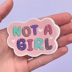 May include: A pink sticker with the words "NOT A GIRL" in bubble letters. The letters are various colors, including teal, purple, and brown. The sticker has a cloud-like border with small white hearts.