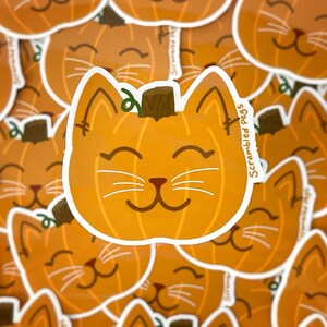 May include: Orange pumpkin-shaped cat sticker with a brown stem and white whiskers. The sticker has the text "Scrambled Pegs" on the side. The cat has a smiling face and is surrounded by other similar stickers.