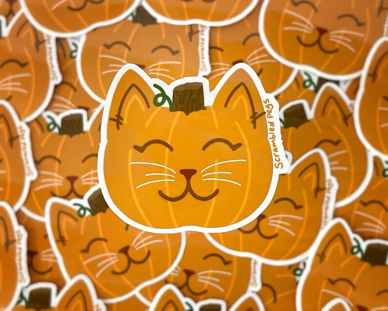 May include: Orange pumpkin-shaped cat sticker with a brown stem and white whiskers. The sticker has the text "Scrambled Pegs" on the side. The cat has a smiling face and is surrounded by other similar stickers.