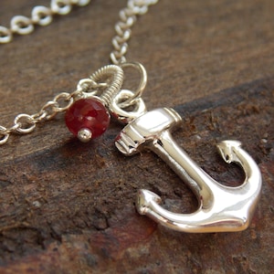 May include: A silver anchor charm necklace with a red gemstone. The necklace is on a wooden surface.