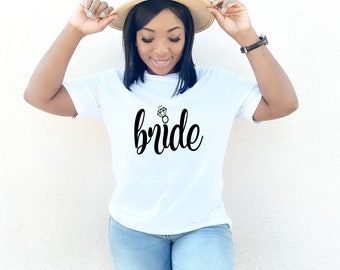 Puede incluir: Una camiseta blanca con la palabra "bride" en escritura negra y un gráfico de anillo de diamantes. La persona lleva un sombrero de paja con una banda negra y vaqueros azules.