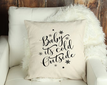 May include: A square, off-white pillow with the phrase "Baby it's cold Outside" in black script. The design includes snowflake and star accents. The pillow is placed on a white chair with a fluffy, white throw.