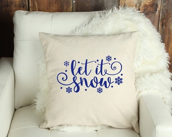 May include: A square, cream-colored pillow with the phrase "let it snow" in a dark blue script. The text is surrounded by blue snowflake designs. The pillow is resting on a white chair with a fluffy white throw.