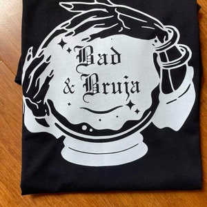 May include: Black t-shirt featuring a white graphic of a crystal ball, hands, and the words "Bad & Bruja" in a gothic font. The design includes stars and a mystical theme. The shirt is folded on a wooden surface.