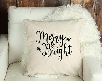 Puede incluir: Un cojín cuadrado de color crema con las palabras "Merry & Bright" en escritura negra. El cojín está sobre una silla blanca con una manta de piel sintética blanca y esponjosa. El cojín tiene un tema festivo y navideño.