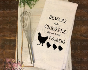 May include: A white kitchen towel with the text "BEWARE of the CHICKENS they can be real PECKERS" and a black chicken and chick silhouette design. A stainless steel whisk is next to the towel.