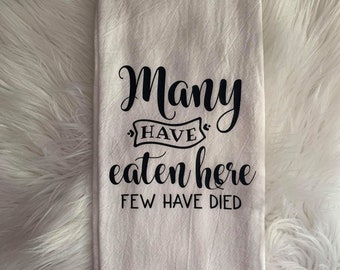May include: Off-white kitchen towel with the text "Many have eaten here, few have died" in black script. The towel is folded and has a textured appearance. The background is a fluffy, white surface.