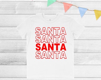 May include: White t-shirt featuring the word "SANTA" in red, outlined letters, repeated four times. The third "SANTA" is solid red. The shirt is on a white wooden surface, with colourful pennant flags strung across the top.