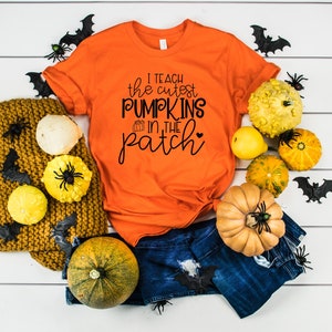 May include: Orange t-shirt with the text "I teach the cutest pumpkins in the patch" and a heart. The shirt is styled with blue jeans, decorative pumpkins, a mustard-colored knit blanket, and Halloween decorations.