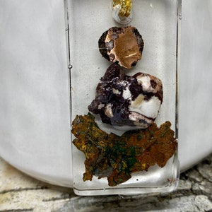 May include: A rectangular pendant with a gold chain, featuring a layered design of three different types of stones. The top stone is a round, copper-colored stone. The middle stone is a white and brown stone with a purple hue. The bottom stone is a large, flat, copper-colored stone with green accents.