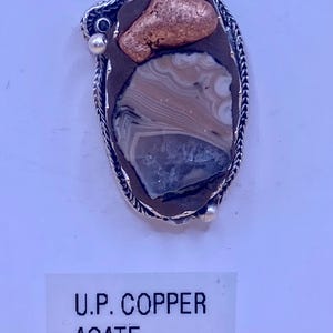 May include: Oval pendant featuring a copper nugget and a polished agate stone, set in a silver-toned frame. The agate displays bands of beige, gray, and white. The text "U.P. COPPER AGATE" is printed on a white label below the pendant.