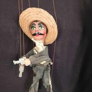 May include: A vintage hand puppet of a man wearing a brown sombrero and a black suit holding a silver pistol. The puppet has a painted face with a mustache and green eyes.