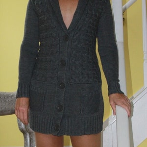 May include: A woman wearing a dark gray, button-down sweater dress with a V-neck and pockets. She is wearing black platform heels. The background is a yellow wall and a white staircase.