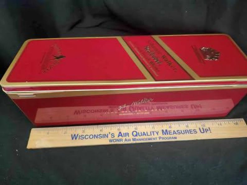 May include: Red metal tin with gold trim and the words "Johnnie Walker" and "Red Label" printed on the front. The tin is closed and has a gold hinge.