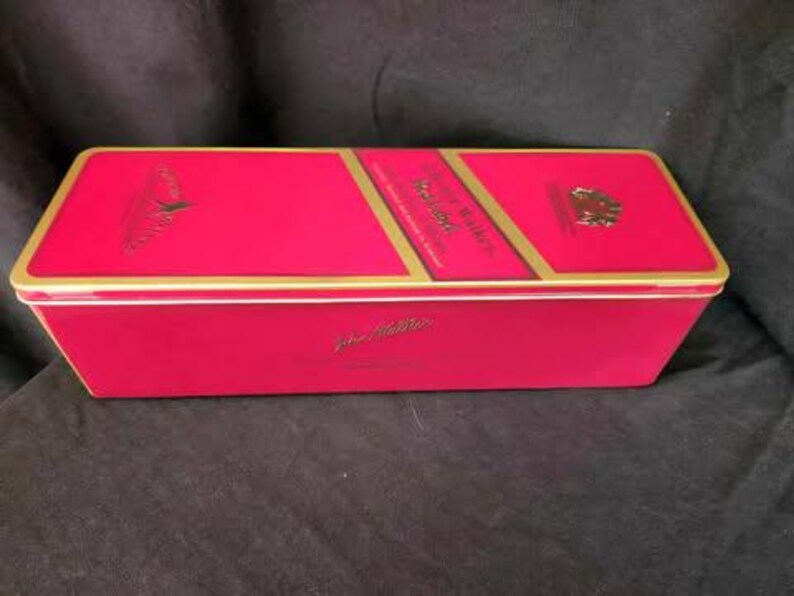 May include: A red and gold tin box with the words "Johnnie Walker" printed on the top. The box is closed and has a gold trim.