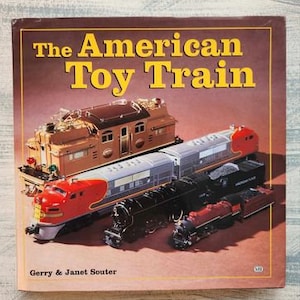 May include: A book titled "The American Toy Train" featuring a toy train set on the cover. The set includes a red, silver, and black locomotive, a brown and gold engine, and a black and red caboose.